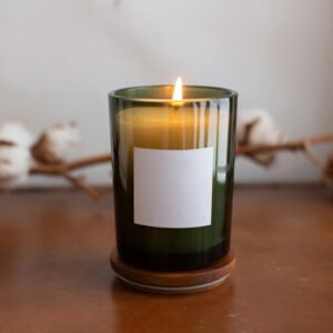 A lit candle in a green glass jar with a blank label, set against a cozy cotton branch backdrop.